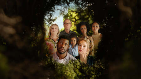 In einer grünen Hecke ist ein Loch, durch das man einen Blick hinter die Hecke werfen kann. Dort ist eine Gruppe von Menschen zu sehen: Ganz vorne die Köpfe von Dawit (Akeem van Flodrop, 2.v.l.) und Laura (Sina Martens, 3.v.r.). Hinter ihnen stehen Heidi (Gabriela Maria Schmeide, l.), Walter (Peter Lohmeyer, 3.v.l.), Fiyori (Lousia Heinricht, M.), Lemlem (Dennenesch Zoudé, 2.v.r.) und Armana (Chioma Chukwuani, r.).