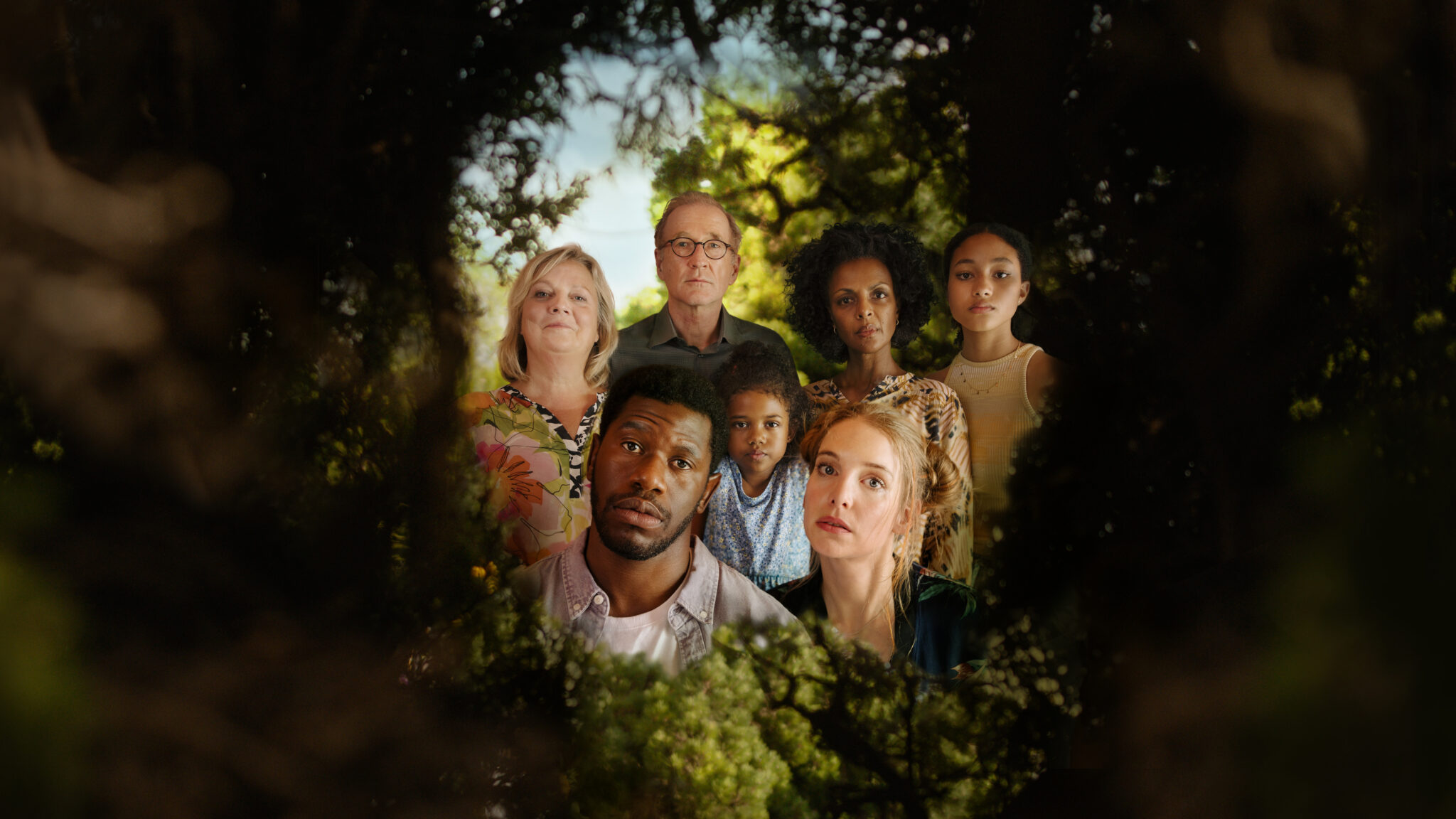 In einer grünen Hecke ist ein Loch, durch das man einen Blick hinter die Hecke werfen kann. Dort ist eine Gruppe von Menschen zu sehen: Ganz vorne die Köpfe von Dawit (Akeem van Flodrop, 2.v.l.) und Laura (Sina Martens, 3.v.r.). Hinter ihnen stehen Heidi (Gabriela Maria Schmeide, l.), Walter (Peter Lohmeyer, 3.v.l.), Fiyori (Lousia Heinricht, M.), Lemlem (Dennenesch Zoudé, 2.v.r.) und Armana (Chioma Chukwuani, r.).