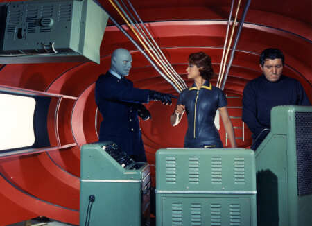 "Fantomas": Fantomas (Jean Marais), Lady Beltham (Marie-Hélène Arnaud) und sein Leibwächter (Henri Attal) stehen in einem roten Raum vor einigen technischen Geräten. Während Fantomas energisch auf ein Gerät zeigt, schaut Lady Beltham ihn direkt an und der Leibwächter schaut konzentriert nach unten.