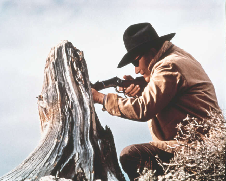 "Der Marshal": Seitenansicht von Rooster Cogburn (John Wayne), der an einem rindenlosen Baumstumpf kniet und mit seinem Gewehr zielt. Er trägt eine derbe Jacke, einen schwarzen Westernhut sowie eine schwarze Augenklappe über dem linken Auge. ‎
