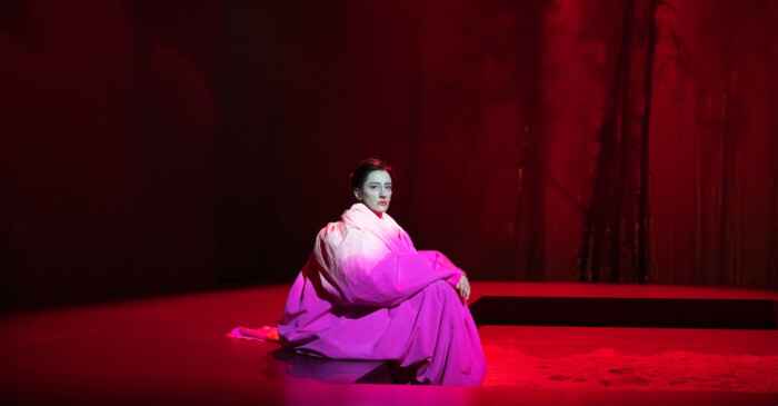 Eine Frau sitzt auf einer Theaterbühne und trägt ein weiß-rosa Kleid.
