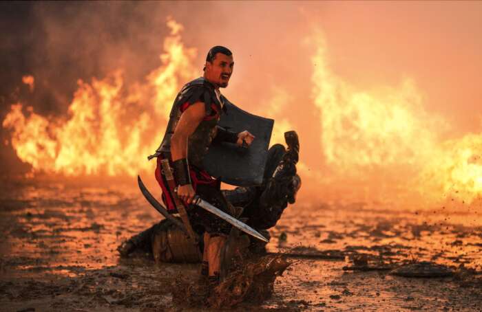Ein Mann mit einem Schwert in der Hand dreht sich um und schreit. Im Hintergrund sind Flammen.