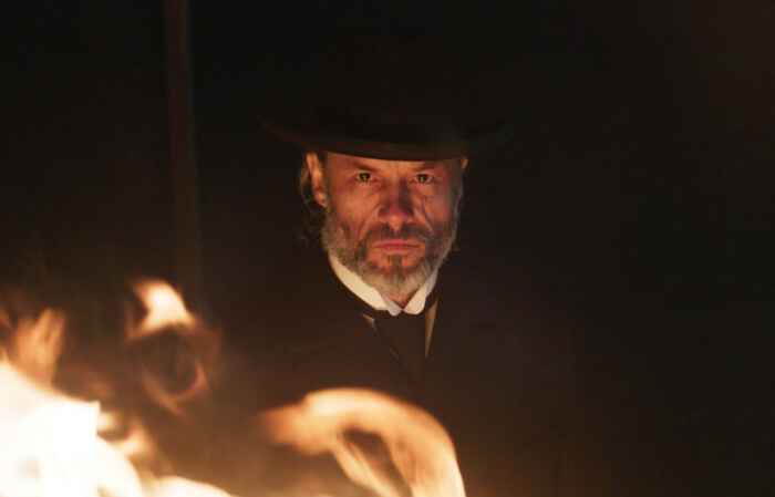 "Brimstone": Der graubärtige Reverend (Guy Pearce) trägt einen schwarzen Anzug und Hut. Er steht in der Dunkelheit und wird nur durch Flammen erhellt. Sein Blick ist stechend.