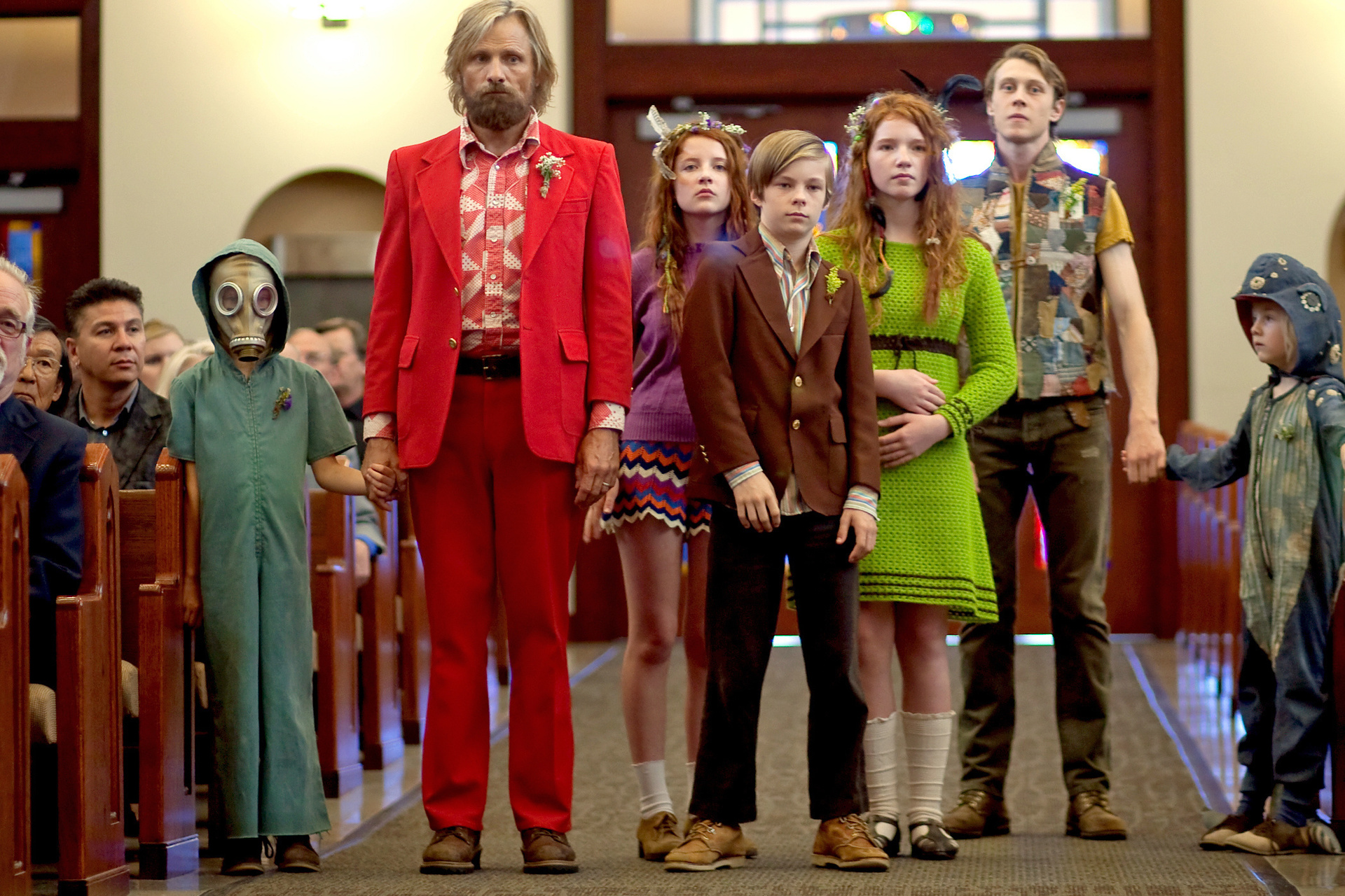 "Captain Fantastic - Einmal Wildnis und zurück": Ben (Viggo Mortensen), Nai (Charlie Shotwell), Bodevan (George MacKay), Kielyr ‎‎(Samantha Isler), Rellian (Nicholas Hamilton), Vespyr (Annalise Basso) ‎und Zaja (Shree Crooks) stehen im Mittelgang eines Kirchenraums und tragen bunte Kleidung.