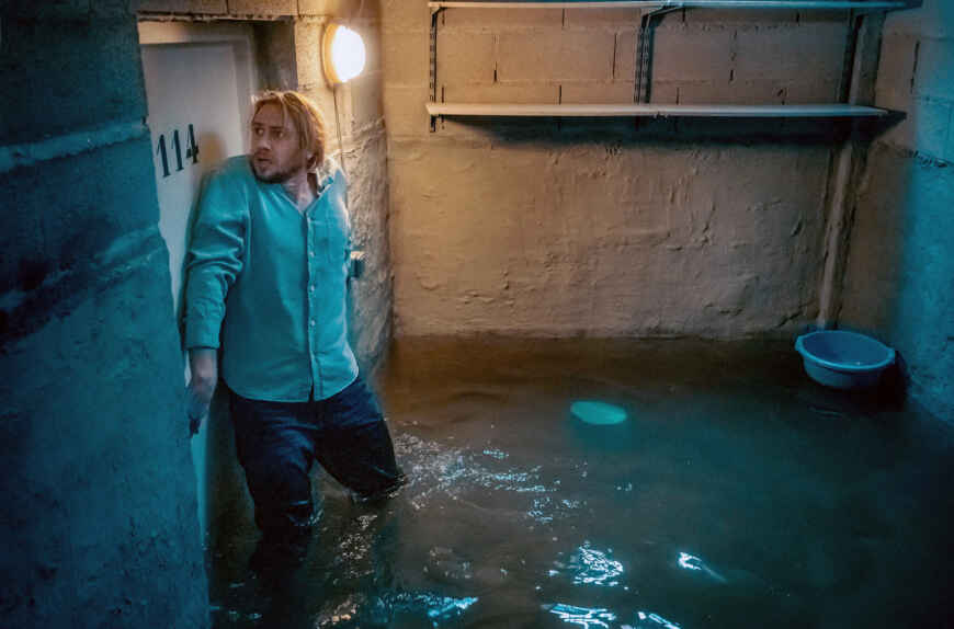 Still aus „Das unsichtbare Wesen“: Ein Mann steht knietief im Wasser in einem Kellerraum. Er drückt seinen Rücken gegen die Tür und versucht, sie mit der rechten Hand zu öffnen.