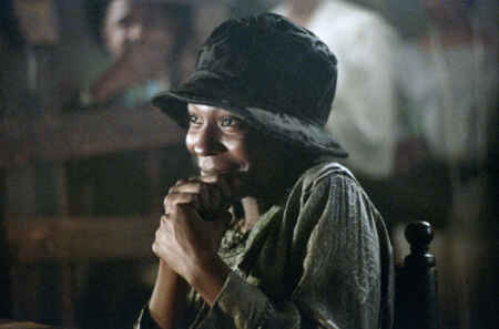 Die erwachsene Celie (Whoopi Goldberg) sitzt lächelnd da und umfasst eine Hand mit der anderen.