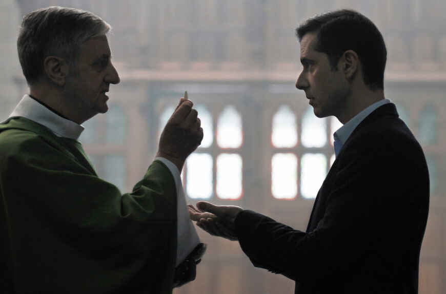 Still aus „Gelobt sei Gott“: Ein Pfarrer mit grünem Gewand hält eine Hostie in die Luft, ihm gegenüber streckt ein Mann im Anzug die Hand aus, um sie zu empfangen.
