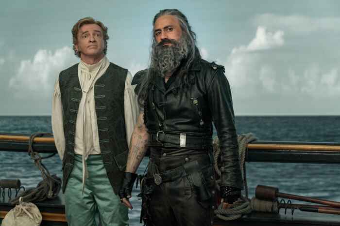 Still aus „Our Flag means Death“: Stede Bonnet (Rhys Darby) und Blackbeard (Taika Waititi) stehen an der Reling eines Schiffes mit dem Rücken zum Meer.