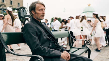 Still aus „Der Rausch“: Martin (Mads Mikkelsen) sitzt auf einer Bank am Hafen und schaut aufs Meer. Im Hintergrund Jugendliche in Matrosenkleidern, die Flaschen mit Alkohol halten.
