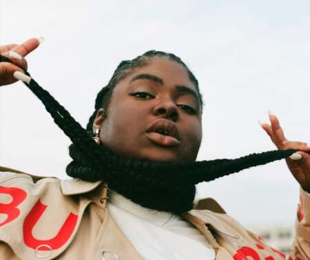 Chika, mit Braids um ihren Hals geschlungen, vor einem hellen Hintergrund.