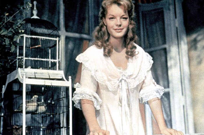 Still aus „Christine“: Christine (Romy Schneider) steht im weißen Kleid auf einem Balkon und lächelt, im Hintergrund ein Vogelkäfig.