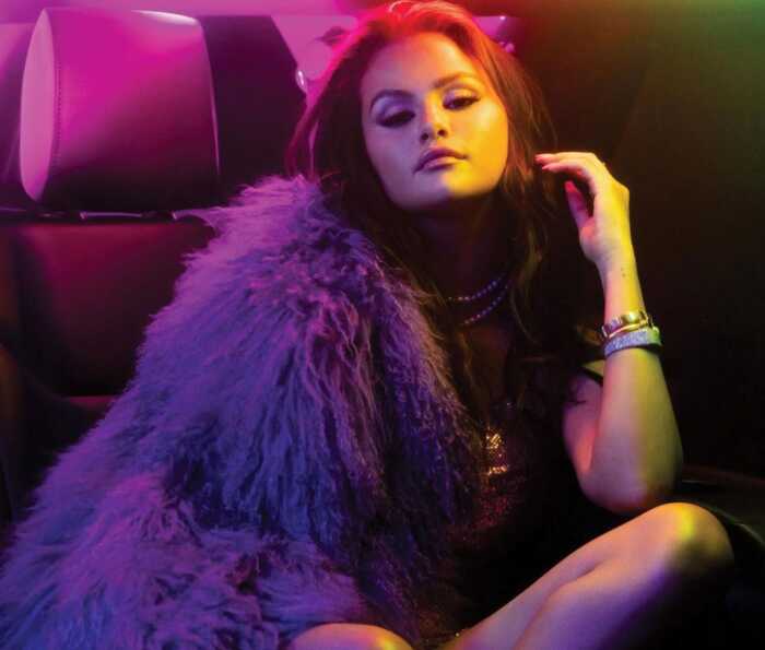 Selena Gomez, sitzend in einem Auto. Das Bild ist in Lilatönen gehalten.