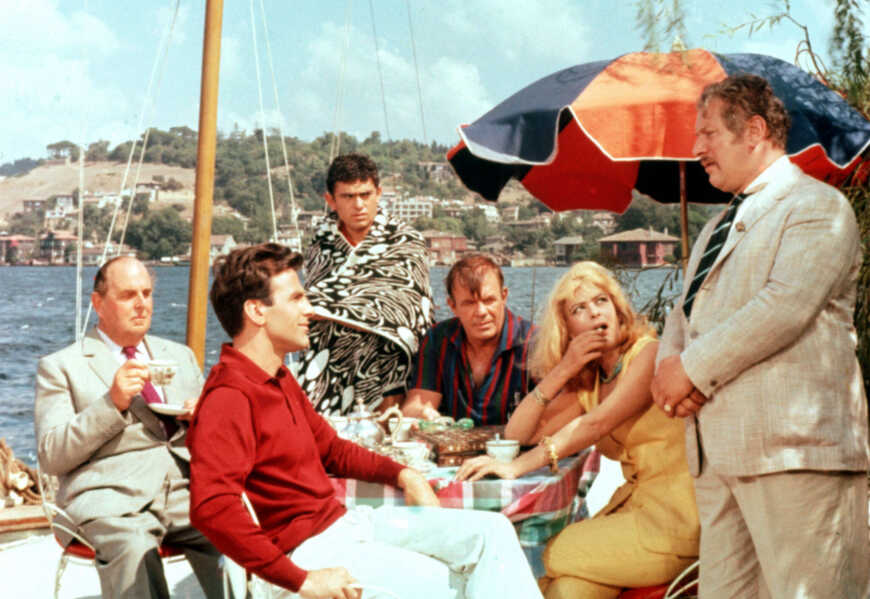 Still aus „Topkapi“: Fünf Menschen sitzen im Freien um einen Tisch herum, im Hintergrund das Meer. Ein sechster Mann steht rechts im Bild vor einem Sonnenschirm.