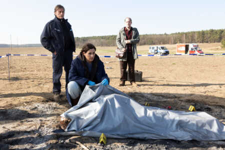 "Das Mädchen von früher": Eine abgedeckte Leiche liegt auf einem trockenen Feld. Kriminalhauptkommissarin Maria Voss (Nina Kunzendorf) kniet daneben. Ihre Kollegen Lars Klapproth (André Hennicke) und Teo Kromann (Godehard Giese) stehen hinter ihr.