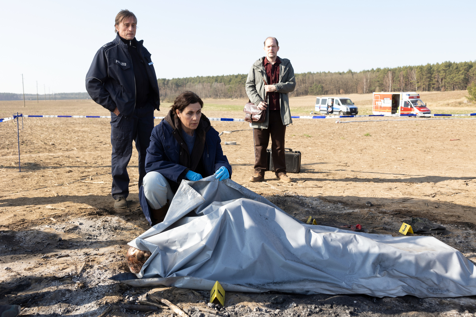 "Das Mädchen von früher": Eine abgedeckte Leiche liegt auf einem trockenen Feld. Kriminalhauptkommissarin Maria Voss (Nina Kunzendorf) kniet daneben. Ihre Kollegen Lars Klapproth (André Hennicke) und Teo Kromann (Godehard Giese) stehen hinter ihr.