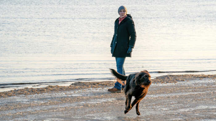 Eine Frau in wetterfester Kleidung geht am Strand entlang. Im Vordergrund läuft ihr Hund auf etwas außerhalb des Bildes zu, sie blickt ihm besorgt hinterher.