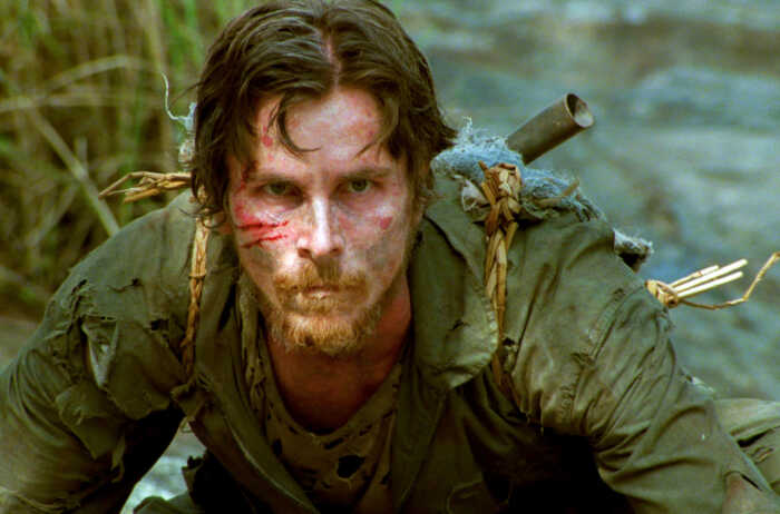 Still aus „Rescue Dawn“: Dieter Dengler (Christian Bale) schaut aus kriechender Position in die Kamera. Er trägt eine zerschlissene Uniform und einen Rucksack aus Stroh und Ästen, Haar und Bart sind struppig, das Gesicht zieren mehrere Wunden.