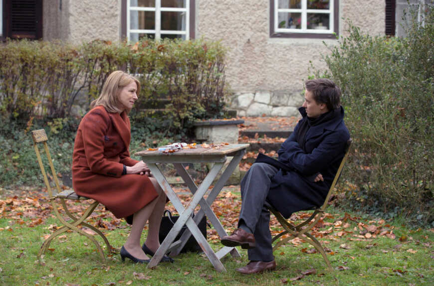 An einem Tisch in einem Hintergarten sitzen eine Frau und ein Mann einander gegenüber. Beide tragen warme Jacken.
