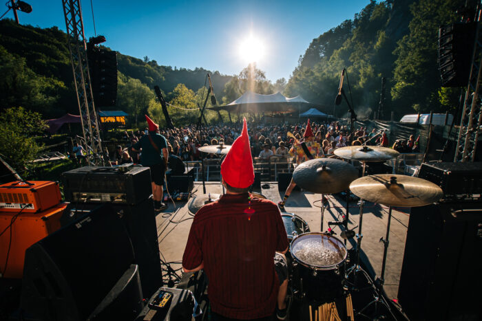 Schlagzeuger spielt auf einer Open-Air Bühne. Vor ihm steht die Menge in schöner Naturkulisse.