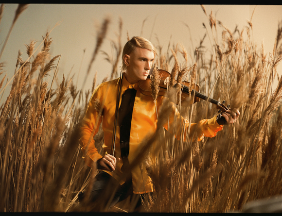 Der Künstler Patrick Wolf steht bei Sonnenuntergang in einem Kornfeld, trägt eine orangefarbene Jacke und spielt auf einer Geige