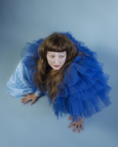 Die Sängerin Alicia Edelweiss trägt ein blaues Kleid und blickt vor blauem Hintergrund schräg zur Seite, während sie sich mit den Händen abstützt.