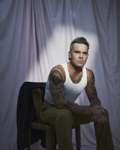 Angestrahlt von einem einzelnen Scheinwerfer, sitzt Robbie Williams im Unterhemd mit gefalteten Händen vor einer Leinwand und guckt missmutig