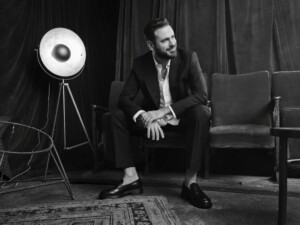 Schwarzweiß-Foto: Der Musiker sitzt auf einer Couch und blickt lächelnd zur Seite, neben ihm steht ein altmodischer Scheinwerfer