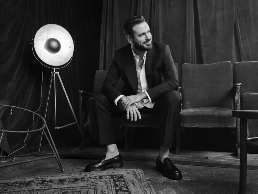 Schwarzweiß-Foto: Der Musiker sitzt auf einer Couch und blickt lächelnd zur Seite, neben ihm steht ein altmodischer Scheinwerfer