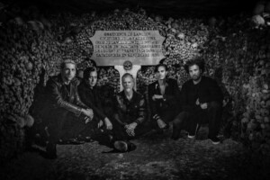 Ein Schwarz-Weiß-Foto zeigt die fünfköpfige Band kniend und sitzend vor einer Wand aus Knochen und Totenschädeln.