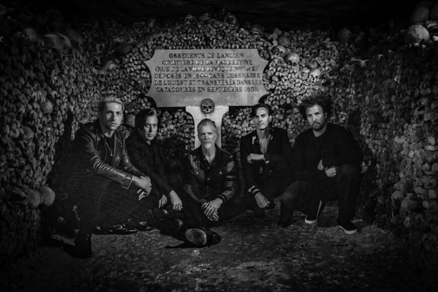 Ein Schwarz-Weiß-Foto zeigt die fünfköpfige Band kniend und sitzend vor einer Wand aus Knochen und Totenschädeln.