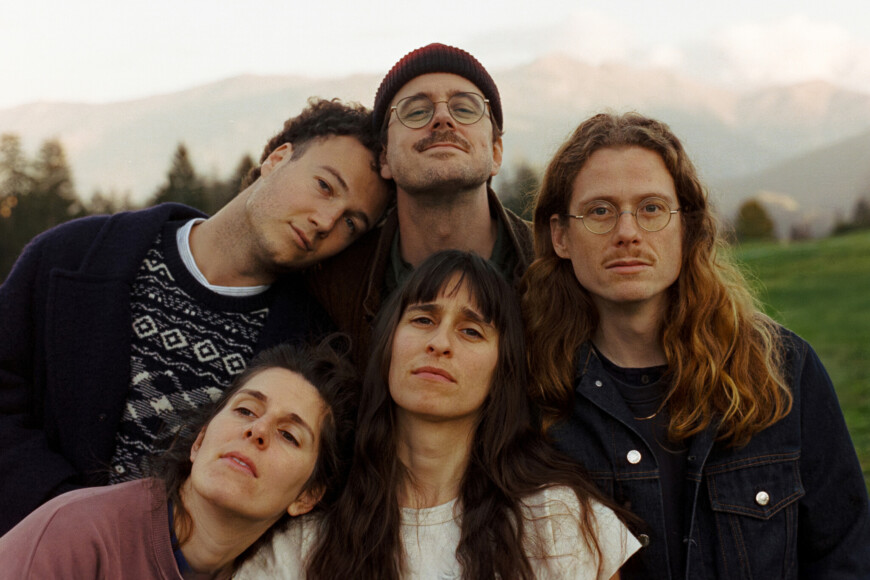 Die Band steht eng zusammen, im Hintergrund Berge.