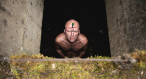 Ein nackter Mann (Rory Kinnear) mit Schnitten am Oberkörper und Gesicht, sowie einem Kleeblatt auf der Stirn, steht in einem quadratischen Loch und schaut Richtung Kamera.