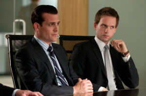 "Suits - Ausgehandelt": Harvey (Gabriel Macht) und Mike (Patrick J. Adams) sitzen nebeneinander an einem Konferenztisch. Beide tragen Anzug und blicken aufmerksam in dieselbe Richtung.