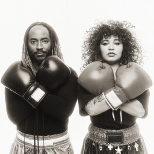 Thundercat und Remi Wolf sind sportlich angezogen, und tragen beide Boxhandschuhe. Sie stehen mit gekreuzten Armen nebeneinander.