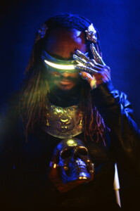 Musiker Thundercat steht im dunkeln mit einer leuchtenden Brille vor der Kamera und hält einen goldenen Totenkopf zu dem er herunter schaut.