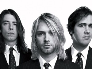 Pressefoto von Nirvana in schwarz-weiß, mit Krist Novoselic (l.), Kurt Cobain (m.) und Dave Grohl (r.). Die Bandmitglieder stehen in Anzügen nebeneinander und schauen in die Kamera.