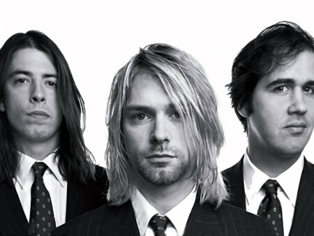 Pressefoto von Nirvana in schwarz-weiß, mit Krist Novoselic (l.), Kurt Cobain (m.) und Dave Grohl (r.). Die Bandmitglieder stehen in Anzügen nebeneinander und schauen in die Kamera.