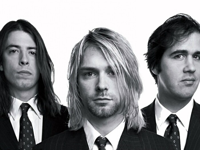 Pressefoto von Nirvana in schwarz-weiß, mit Krist Novoselic (l.), Kurt Cobain (m.) und Dave Grohl (r.). Die Bandmitglieder stehen in Anzügen nebeneinander und schauen in die Kamera.