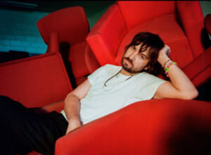 Kevin Parker aka Tame Impala sitzt zwischen roten Sesseln und schaut in die Kamera.