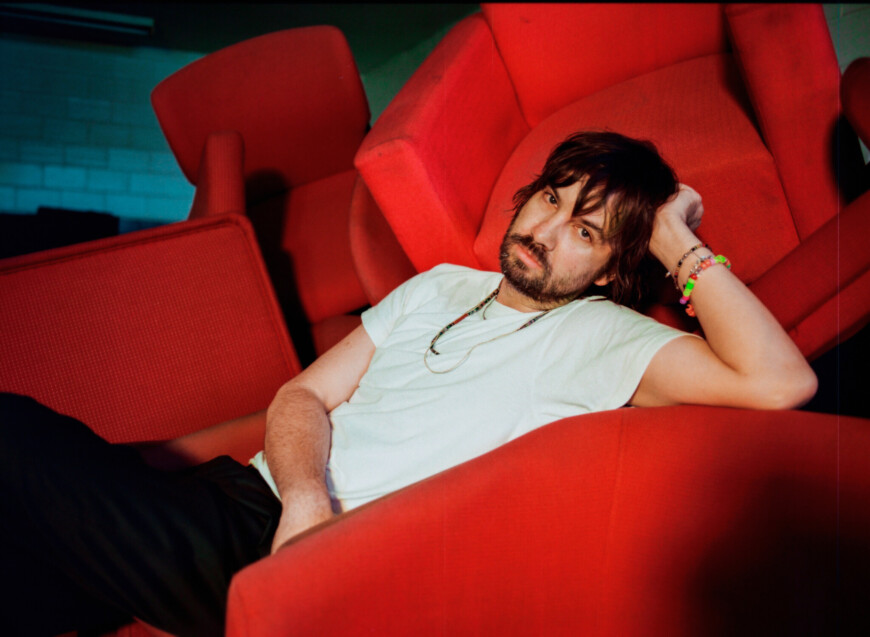 Kevin Parker aka Tame Impala sitzt zwischen roten Sesseln und schaut in die Kamera.
