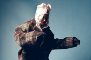 HAAi steht dynamisch/tanzend vor einem blauen Hintergrund. Sie trägt eine Wollmütze und eine karierte Winterjacke.