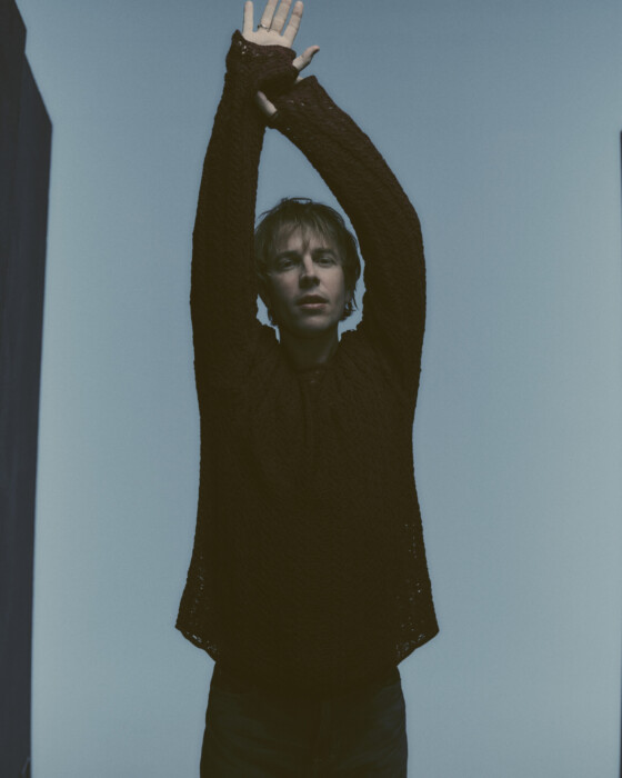 Tom Odell steht vor einem blauen Hintergrund und streckt beide Arme in die Luft.