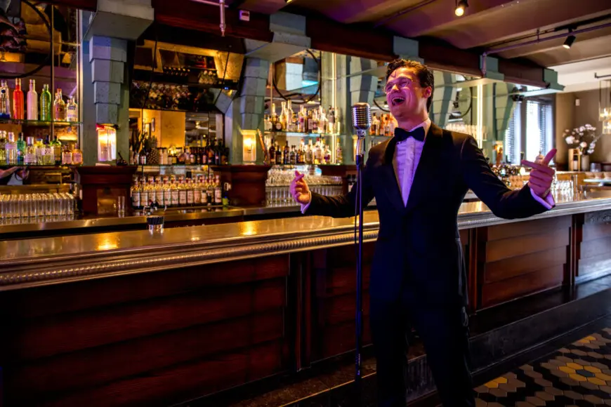 Dennis van Aarssen steht in einem Anzug in einer beleuchteten Bar an einem Mikrofon und singt.