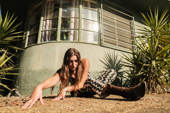 Marla Moya sitzt auf sandigem Boden vor einem grünen Haus und Pflanzen. Sie lehnt sich mit den Armen nach vorne, Richtung Kamera.