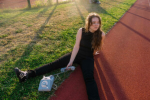 Nina Caroline sitzt im Sonnenlicht auf dem Boden, schaut in die Ferne und hält ein altes Kabeltelefon in der Hand.