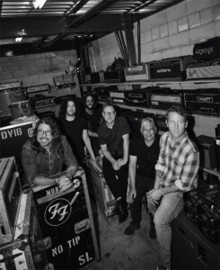Die Band Foo Fighters steht im Backstage zwischen Instrumenten, Boxen und Verstärkern und schaut zur Kamera, das Bild ist in schwarz-weiß.