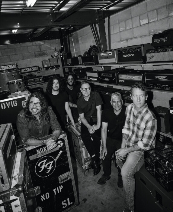 Die Band Foo Fighters steht im Backstage zwischen Instrumenten, Boxen und Verstärkern und schaut zur Kamera, das Bild ist in schwarz-weiß.