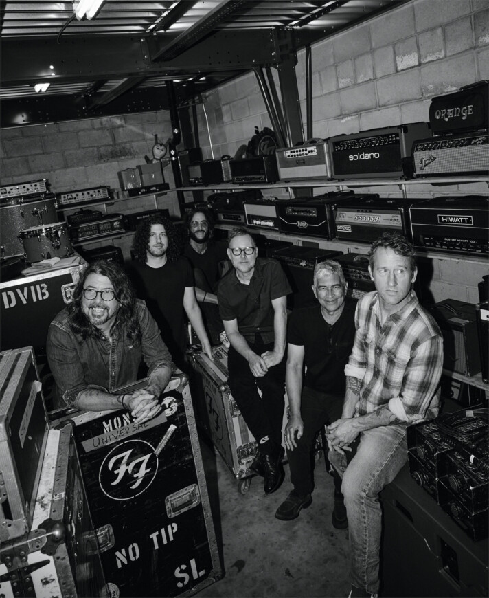 Die Band Foo Fighters steht im Backstage zwischen Instrumenten, Boxen und Verstärkern und schaut zur Kamera, das Bild ist in schwarz-weiß.