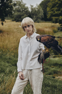 Avalon Emerson steht in einem Feld und trägt einen Greifvogel auf dem Arm.