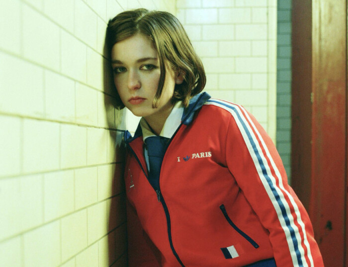 Sängerin Snail Mail lehnt mit dem Kopf an einer Wand aus Fliesen und schaut neutral in die Kamera. Das Bild hat einen grünlichen Filter.